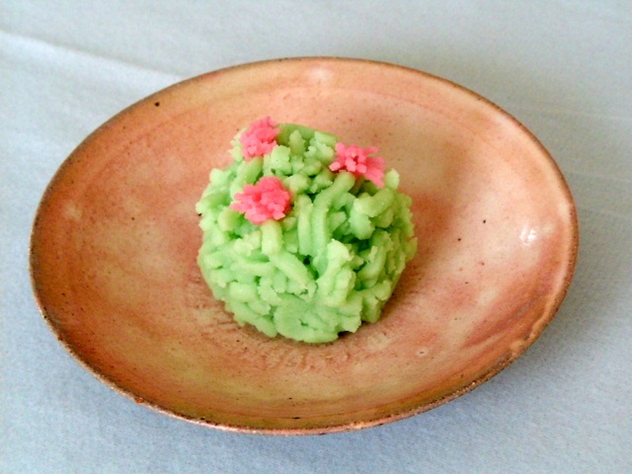 Green mound-shaped confection with pink flower decorations on a brown ceramic plate, photographed on a light blue tablecloth.