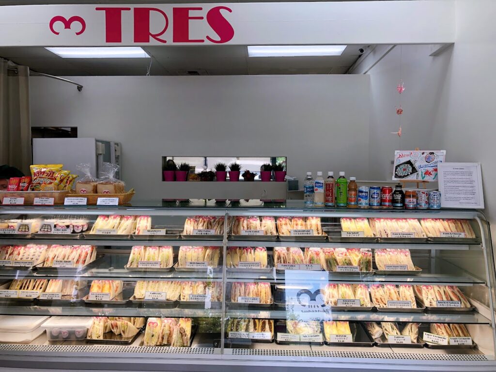 Glass refrigerated display case full of assorted sandwich trays at a cafe counter, with drinks and snacks on top.