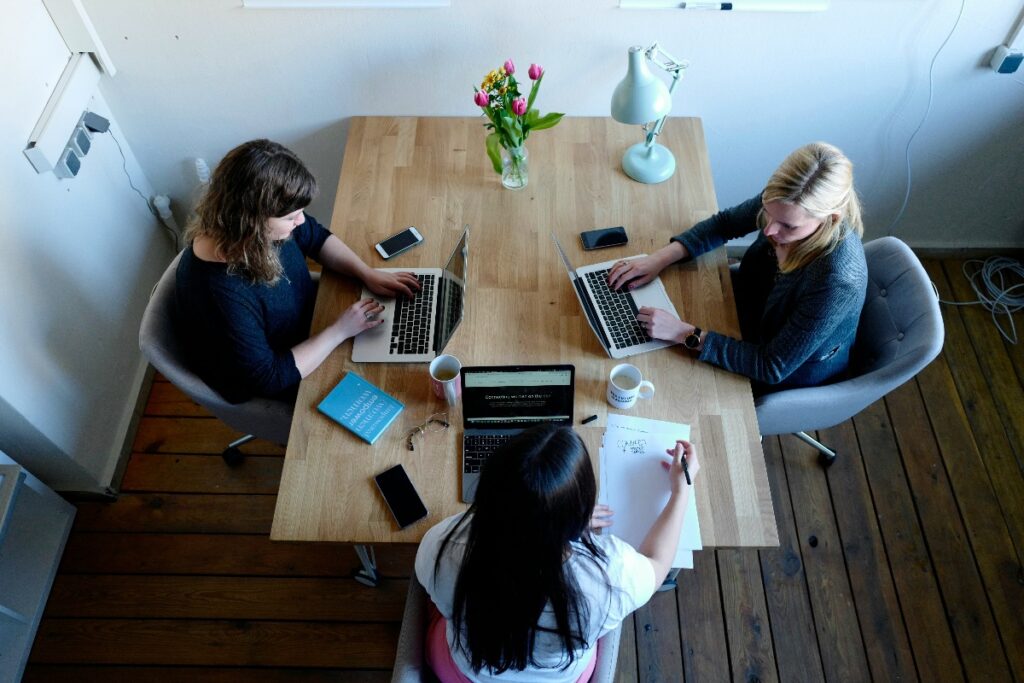 Top-down view of three people around a wooden table working on laptops; phones, notebooks, mugs and a vase of flowers on the table as they collaborate.