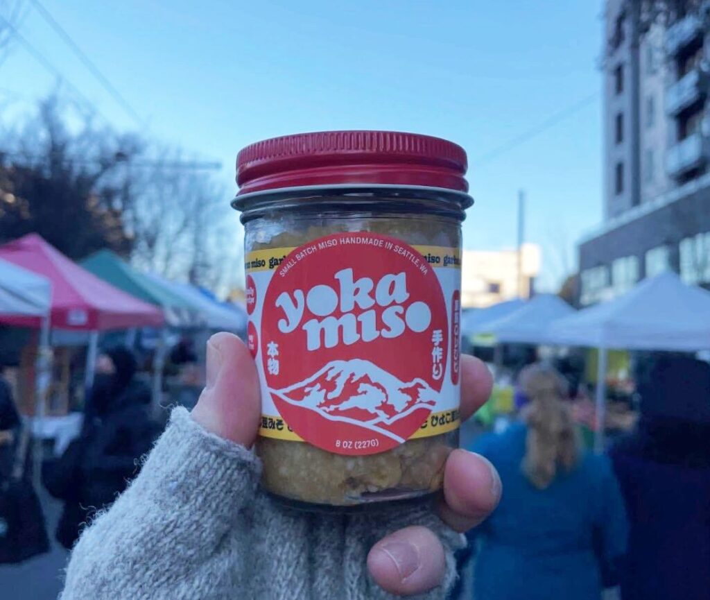 Hand holding a jar of Yokamiso miso with a red label at an outdoor market stall, gloved hand visible in foreground.