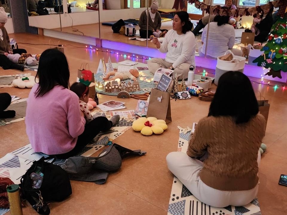 Group of adults and children seated on the floor, making holiday crafts around a festive display in a warmly lit room.
