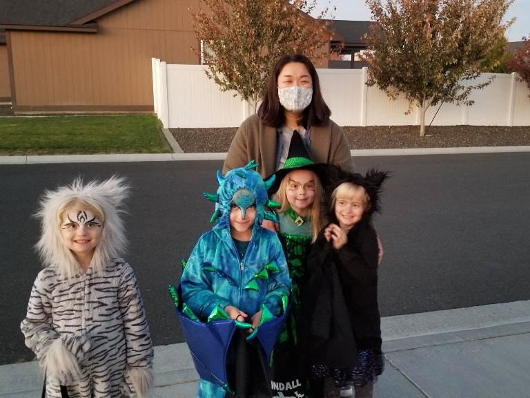 Adult woman with a face mask stands behind four children in Halloween costumes on a sidewalk: a tiger, a blue dragon, a green witch, and a black-clad vampire.