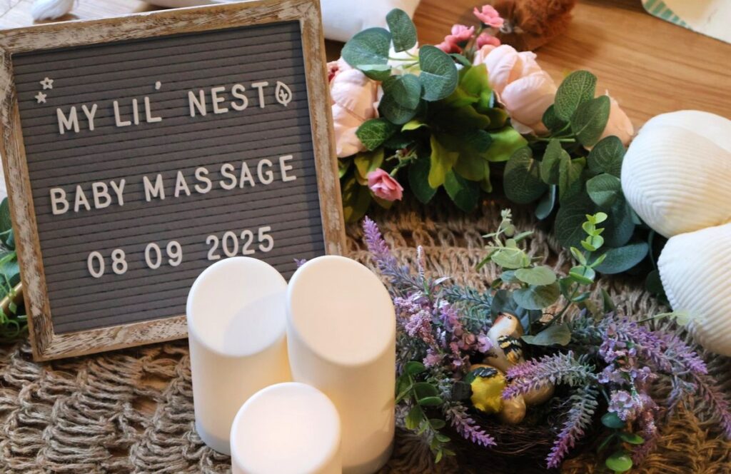 Decorative letter board reading 'My Lil Nest Baby Massage 08 09 2025' beside white candles and a floral arrangement on a woven mat.
