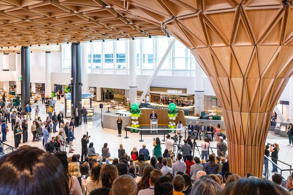 Crowd gathers in a modern atrium as a speaker delivers a speech at a podium with cameras and balloons nearby.