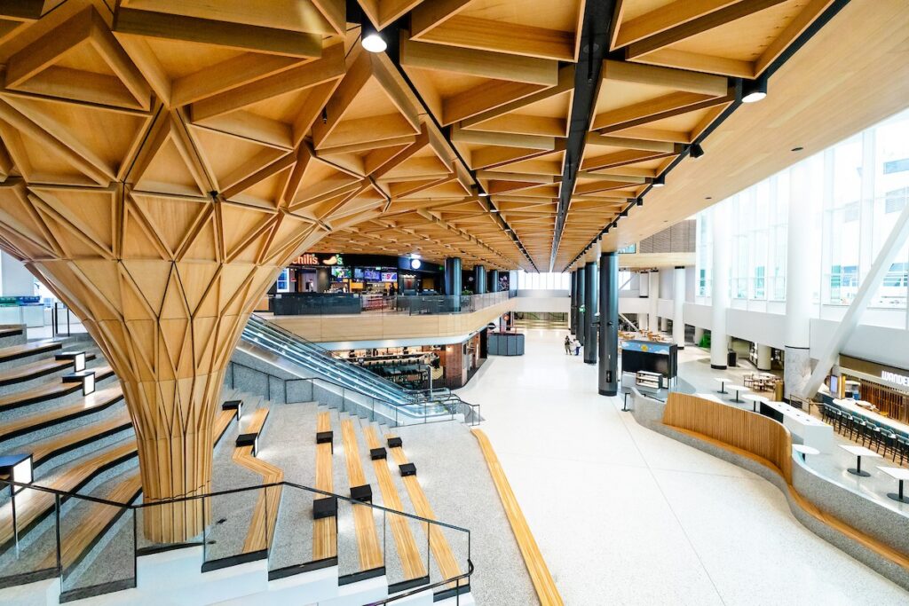 Interior atrium of a modern building with a striking wooden geometric ceiling, tiered seating, and glass railing at the staircase heightening openness.