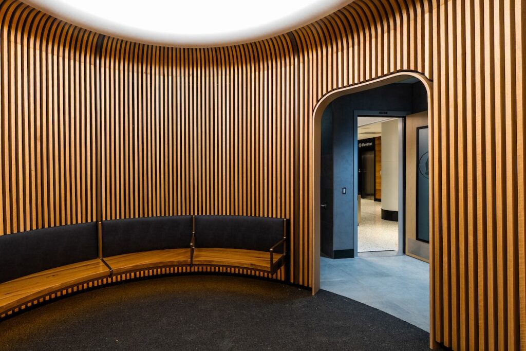 Curved wooden slat wall with a matching curved bench in a modern seating area, an arched doorway opens to a blue-toned hallway.