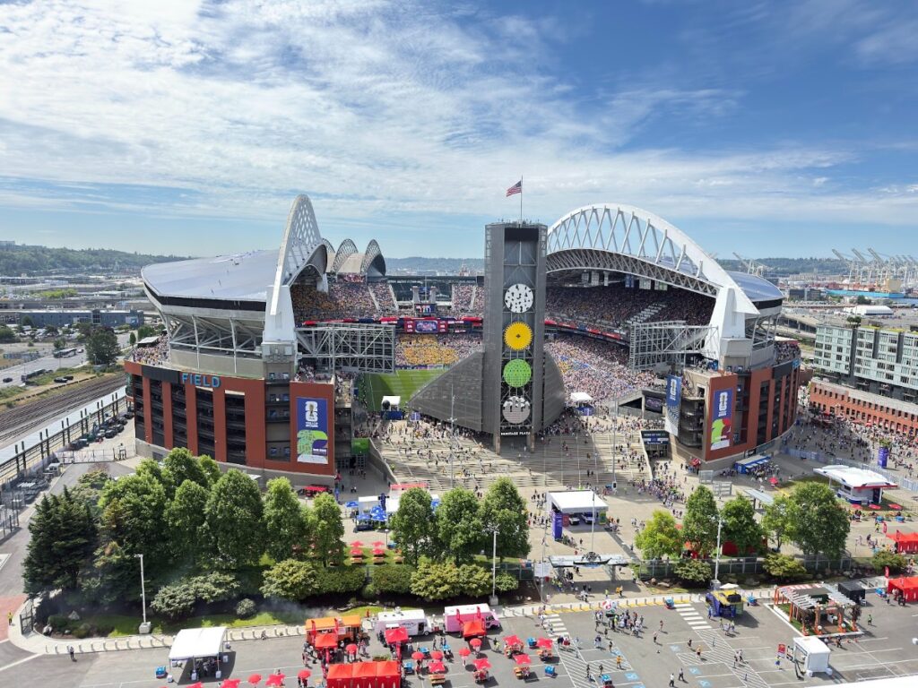 Aerial view of a large modern stadium filled with spectators, featuring a tall central tower with a clock and colored discs, and festival tents outside nearby.