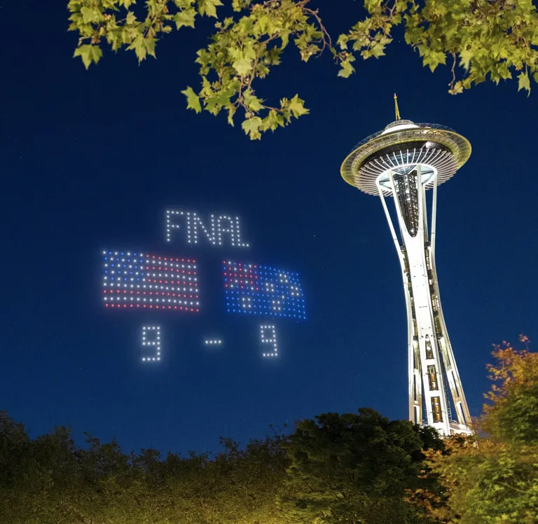 Night shot of the Space Needle at dusk with a lit scoreboard reading FINAL and two US flags, score 9-9.