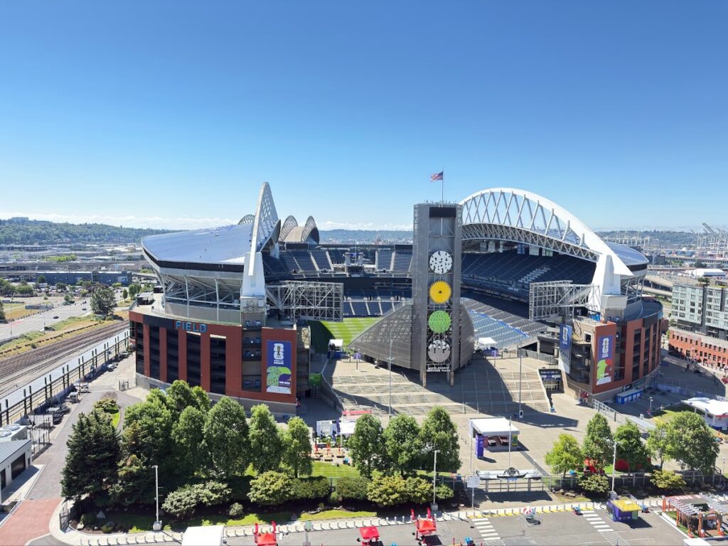 fifa-lumen-field-2026-1 - junglecity.com Aerial view of a large modern stadium with white arches, a tall clock-tower displaying colored discs, and a green field below under a clear blue sky.