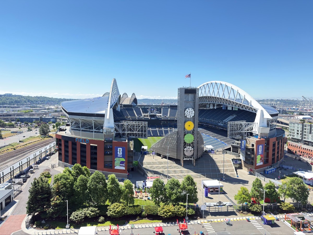Aerial view of a large modern stadium with white arches, a tall clock-tower displaying colored discs, and a green field below under a clear blue sky.