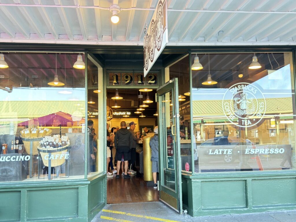 first-starbucks-10 - junglecity.com Open green storefront cafe with the door propped open; people queue inside under warm hanging lights, Starbucks branding on the window.