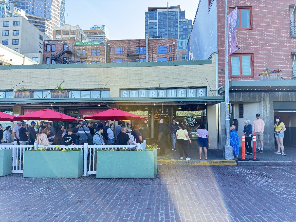 Crowd of people outside a Starbucks on a brick-paved street under red umbrellas and planters in front of the shop with a city backdrop.