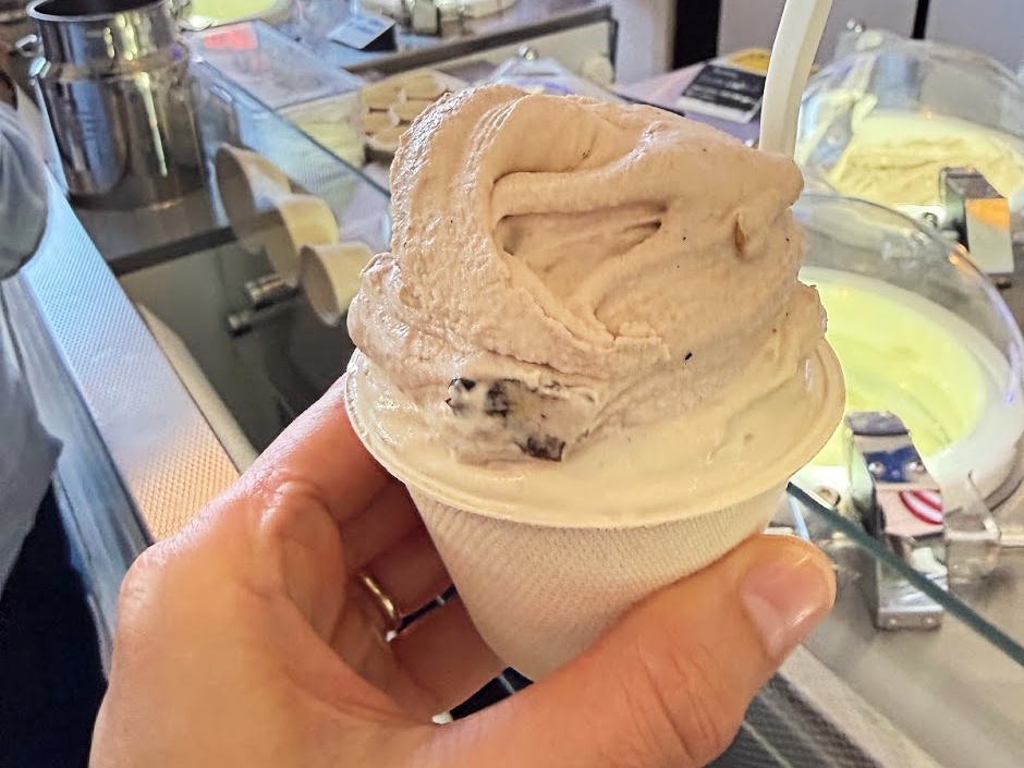 Soft-serve ice cream in a waffle cone with light brown swirls and cookie pieces, held up at a shop counter.