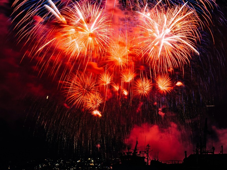 Fireworks explode in red and orange above a dark harbor, with silhouetted ships on the water and city lights below.