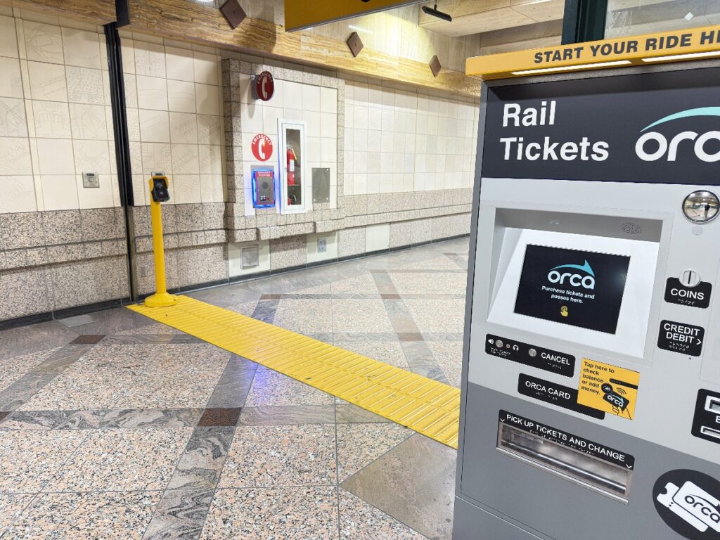 light-rail-15 - junglecity.com Rail ticket vending machine labeled ORCA on the right in a station lobby with a bright yellow tactile guidance path leading toward the wall and emergency equipment in the background.
