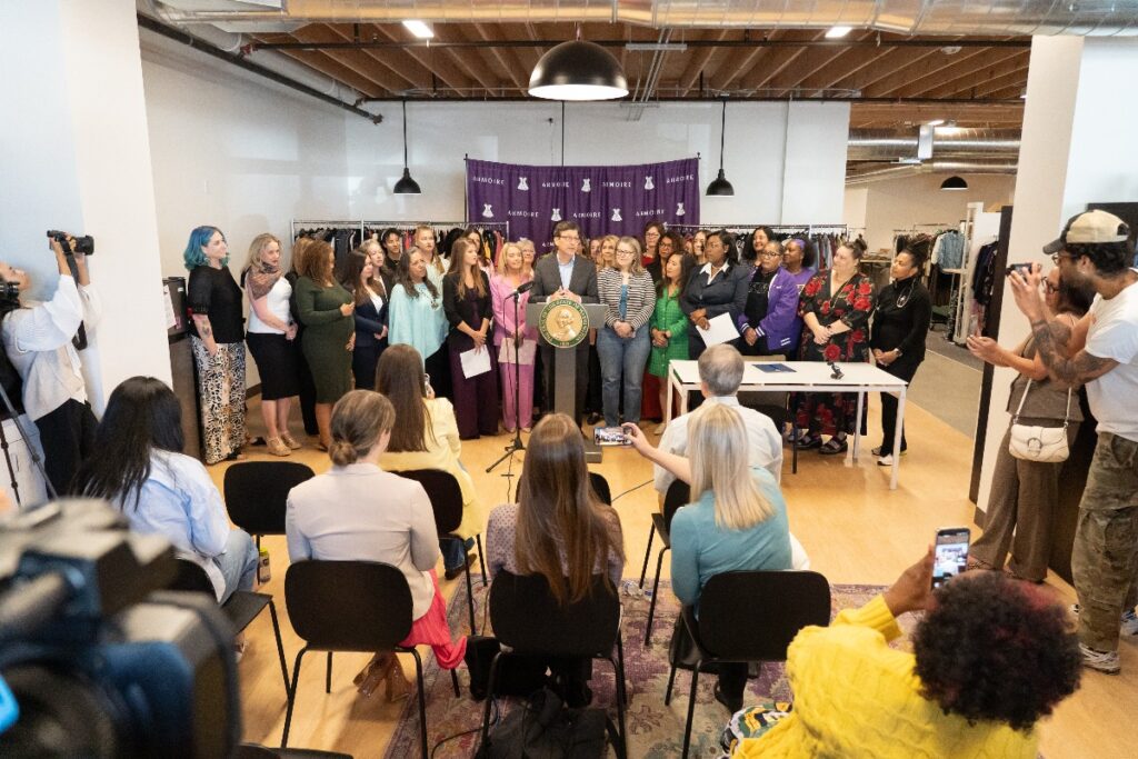 Diverse group stands behind a podium with a seal while an audience sits and records in a bright indoor event space.