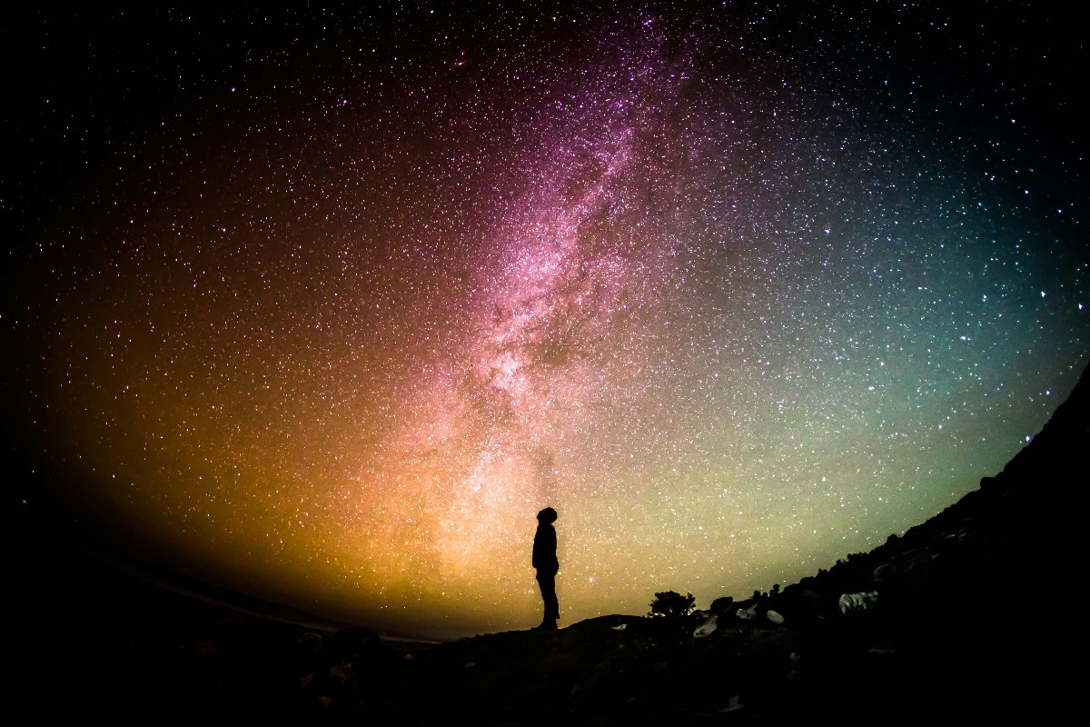 Silhouette of a person standing on a hill under a colorful, star-filled Milky Way night sky for a dramatic landscape.