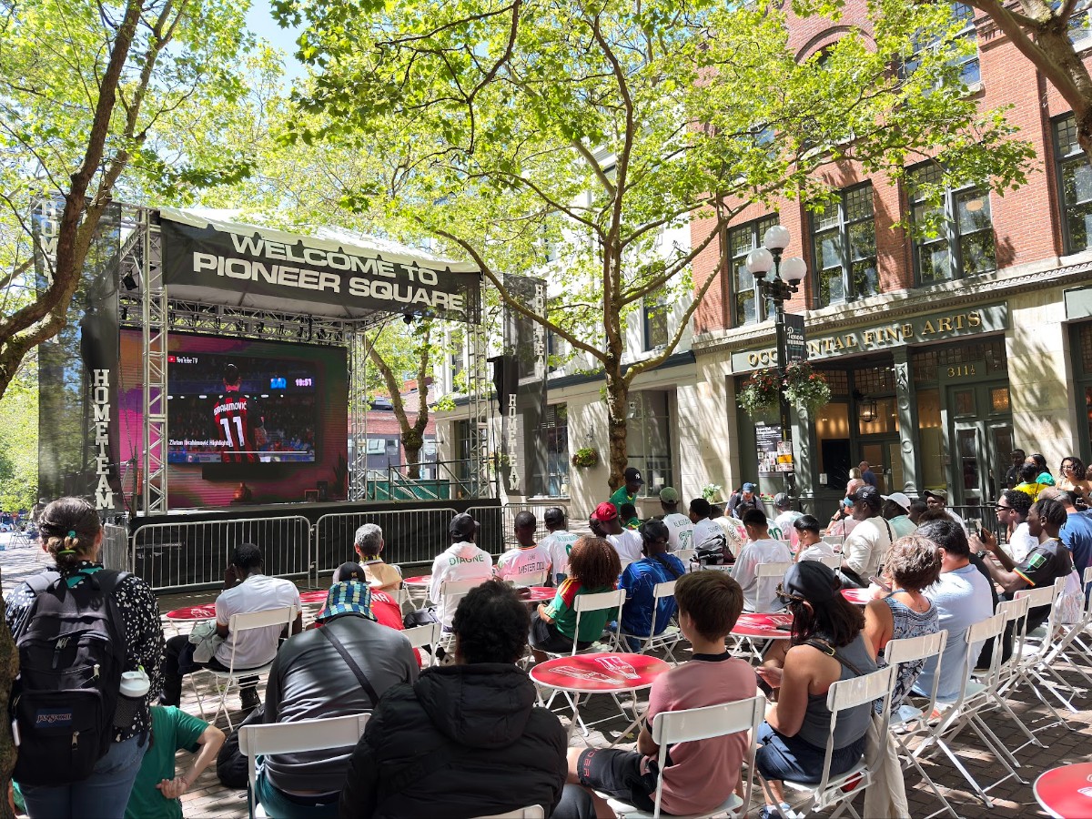 Outdoor crowd watching a large screen on a stage labeled 'Welcome to Pioneer Square' under green trees in a city plaza.