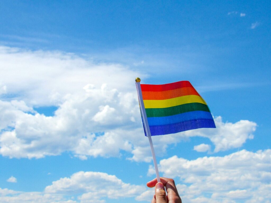 pride-flag-1 - junglecity.com Person holding a rainbow Pride flag against a blue sky with clouds.