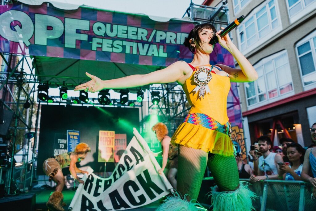 Performer in a bright yellow outfit sings into a microphone on a Pride festival stage with a large colorful Pride banner behind her.