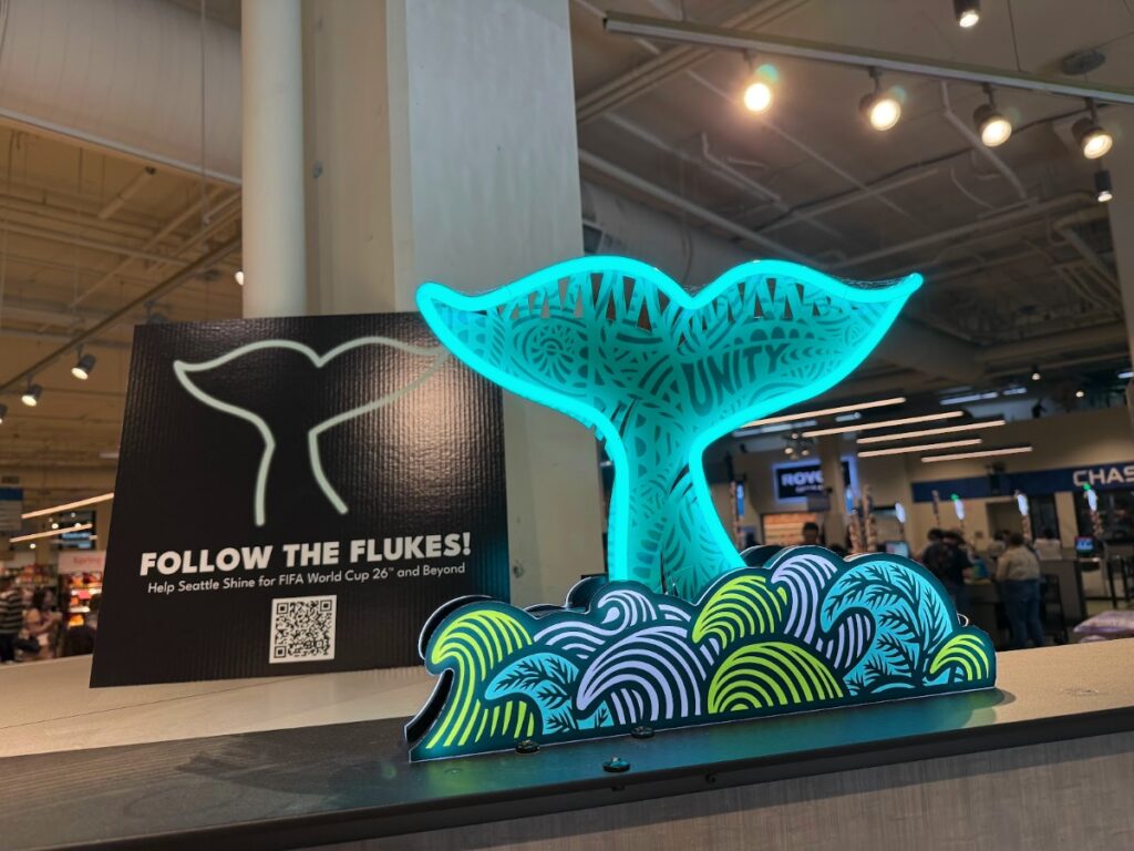 sea-beacon-4 - junglecity.com Neon turquoise whale-tail sculpture with intricate cutout pattern sits above a colorful wave display in an indoor setting.