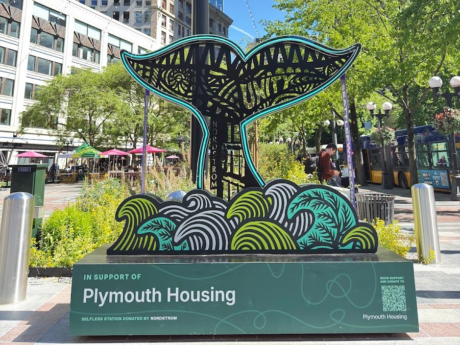 Whale-tail sculpture with teal outlines rising above stylized waves in a city plaza; base reads 'In Support of Plymouth Housing'.