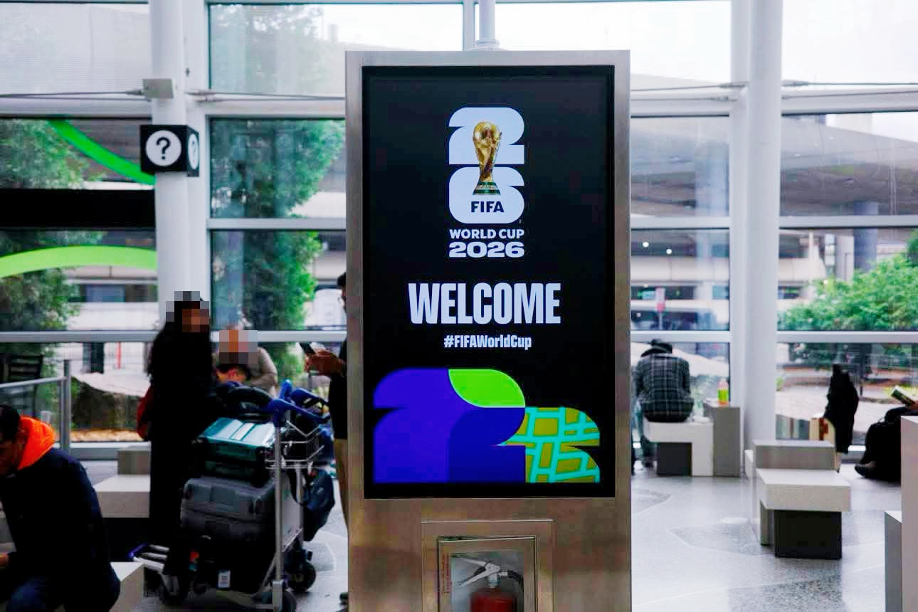 Digital welcome sign for FIFA World Cup 2026 with the trophy logo and the word 'WELCOME' in a busy airport-style area. "#FIFAWorldCup" appears below the message, with travelers and luggage nearby.