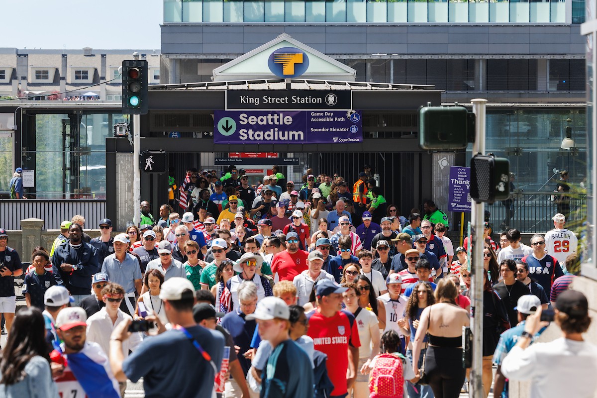 Crowd of fans walking toward the Seattle Stadium entrance under a King Street Station sign, many wearing team jerseys and hats in bright daylight.
