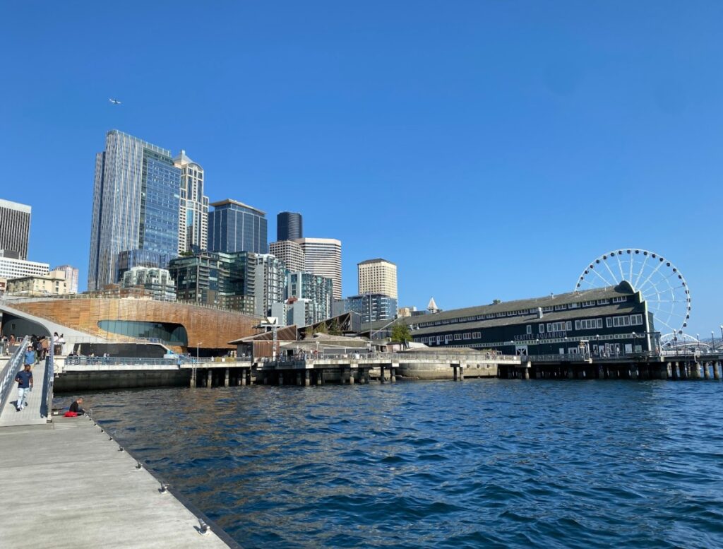 Seattle waterfront skyline with tall buildings, a curved brick pavilion, and a large Ferris wheel by the pier.