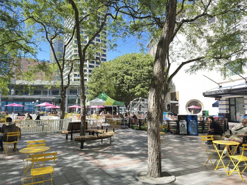 westlake-park-1 - junglecity.com Outdoor plaza with yellow tables and chairs under leafy trees, people sitting and a green tent in the background.