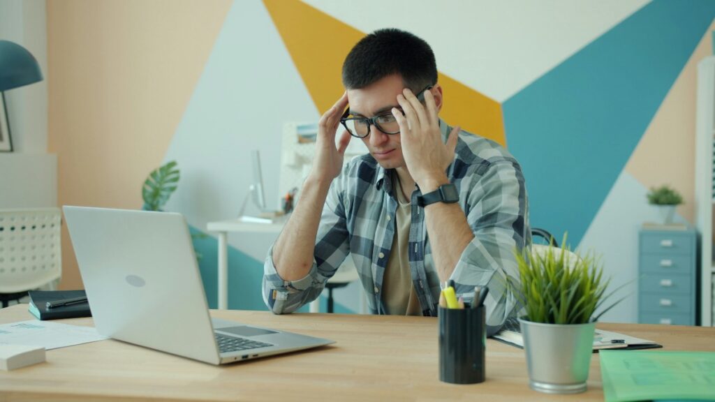 Man wearing glasses sits at a desk, holding his temples in frustration while looking at a laptop.