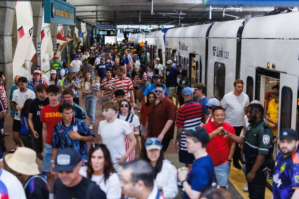 Crowded subway platform with a silver train on the right and a diverse group of people waiting to board or pass by.