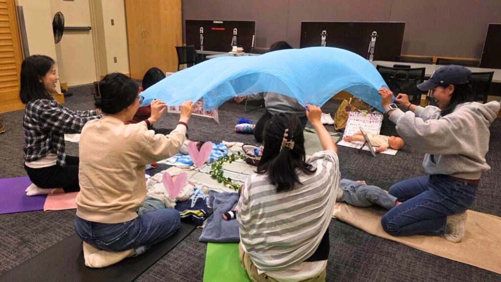 Group of caregivers kneeling on mats, lifting a blue fabric to form a canopy over a baby doll for a care exercise over a classroom setup.