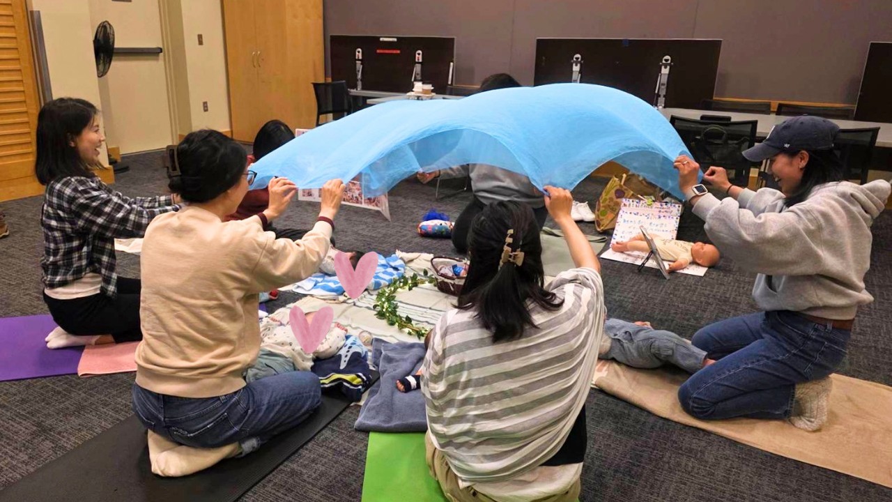 Group of caregivers kneeling on mats, lifting a blue fabric to form a canopy over a baby doll for a care exercise over a classroom setup.