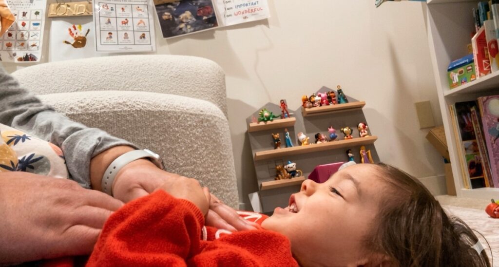 yuko-baby-massage-6 - junglecity.com Child lying on the floor, smiling, as an adult holds their hands; a cozy playroom with toy shelves in the background.
