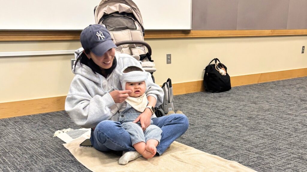 yuko-baby-massage-7 - junglecity.com Woman in a gray hoodie and navy NY cap sits cross‑legged on a beige blanket, smiling at a baby in a helmet indoors beside a stroller.