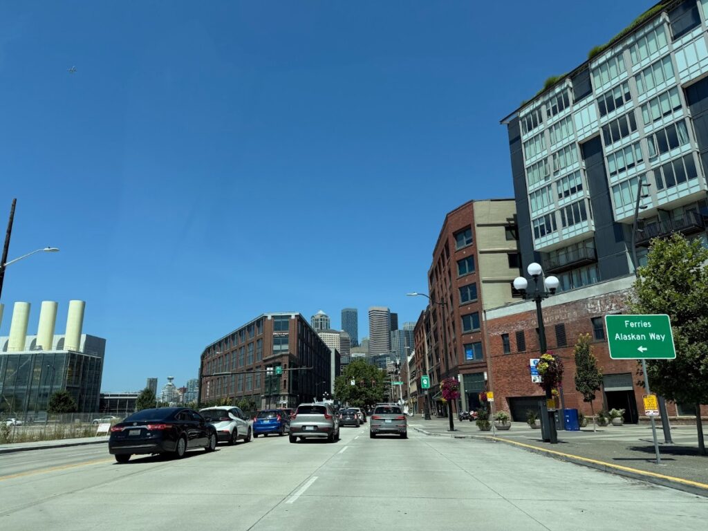 alaskan-way-seattle-1 - junglecity.com Downtown street scene with cars in traffic, brick and glass buildings on the right under a clear blue sky.