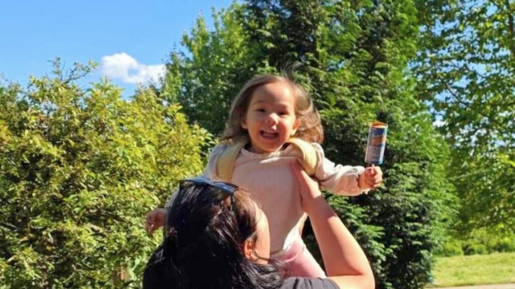 Little girl being held up by an adult, smiling with a blue popsicle in her hand in a sunny park setting.