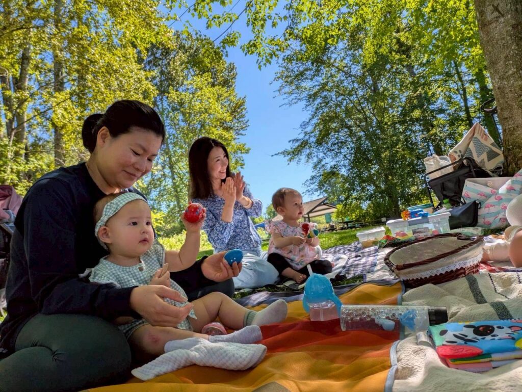 baby-massage-7 - junglecity.com Mother and grandmother watch two babies playing with toys on a sunny park picnic blanket under trees.
