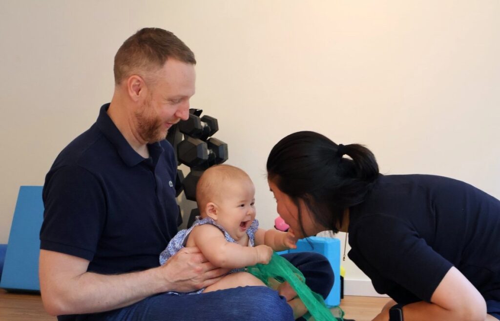 baby-massage-8 - junglecity.com Father holding a laughing baby as a woman leans in, engaging the child during indoor playtime.