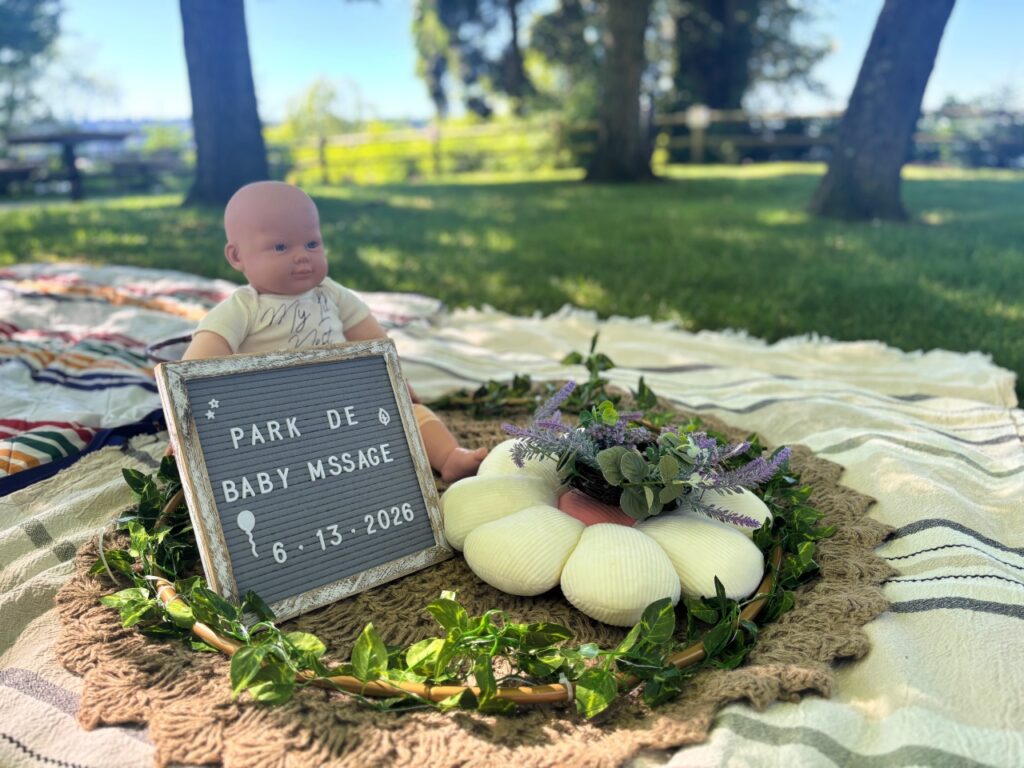 baby-massage-9 - junglecity.com Baby doll seated on a blanket in a sunny park, beside a signboard that reads ‘Park de Baby Message 6-13-2026’ with a shell centerpiece and lavender flowers.