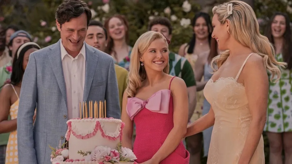 elle-drama-2 - junglecity.com Man in a light blue blazer stands beside a pink-dressed woman and a blonde in cream, smiling at a white cake with pink frosting and candles outdoors.