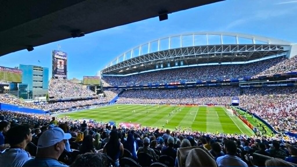 Packed outdoor soccer stadium during daytime with players on the green field and a full crowd in the stands.