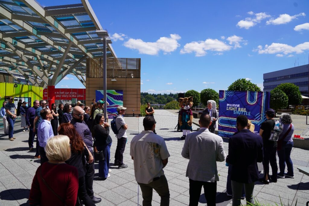 Crowd of people gathered in an outdoor plaza for a Bellevue Light Rail Shuttle event, with a bear mascot on stage and a colorful banner nearby.