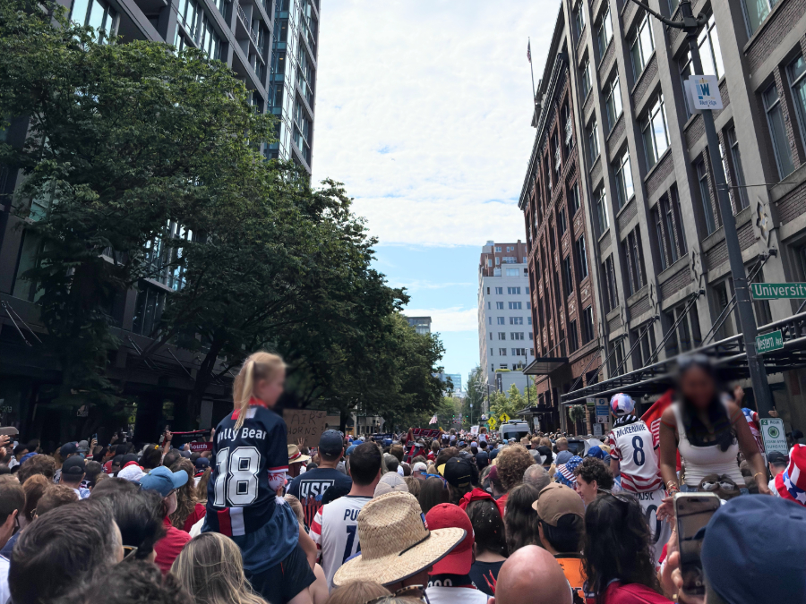 march-to-the-stadium-06-19-2026 - junglecity.com Dense urban street protest with a packed crowd of spectators and participants, tall buildings on both sides, and a girl wearing a black-and-white striped jersey on someone's shoulders (#18).