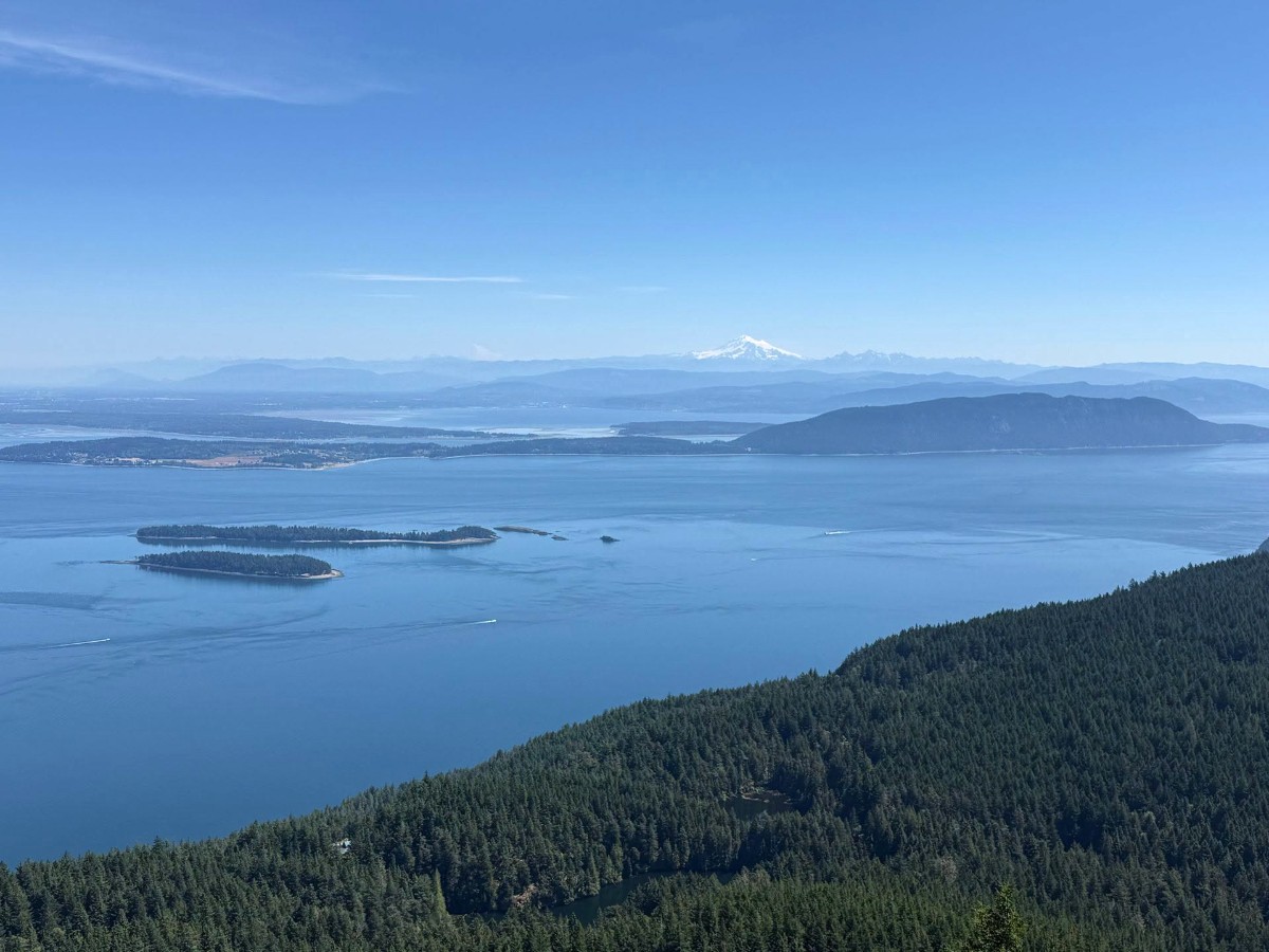 Panoramic blue seascape: islands dotting calm water with a forested slope in the foreground and snow-capped mountains in the distance under a clear sky