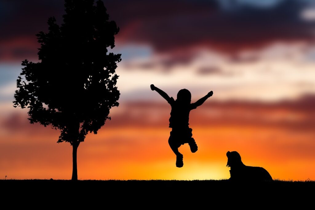 Silhouette of a child jumping with arms raised beside a tree and a resting dog at sunset on a grassy hill