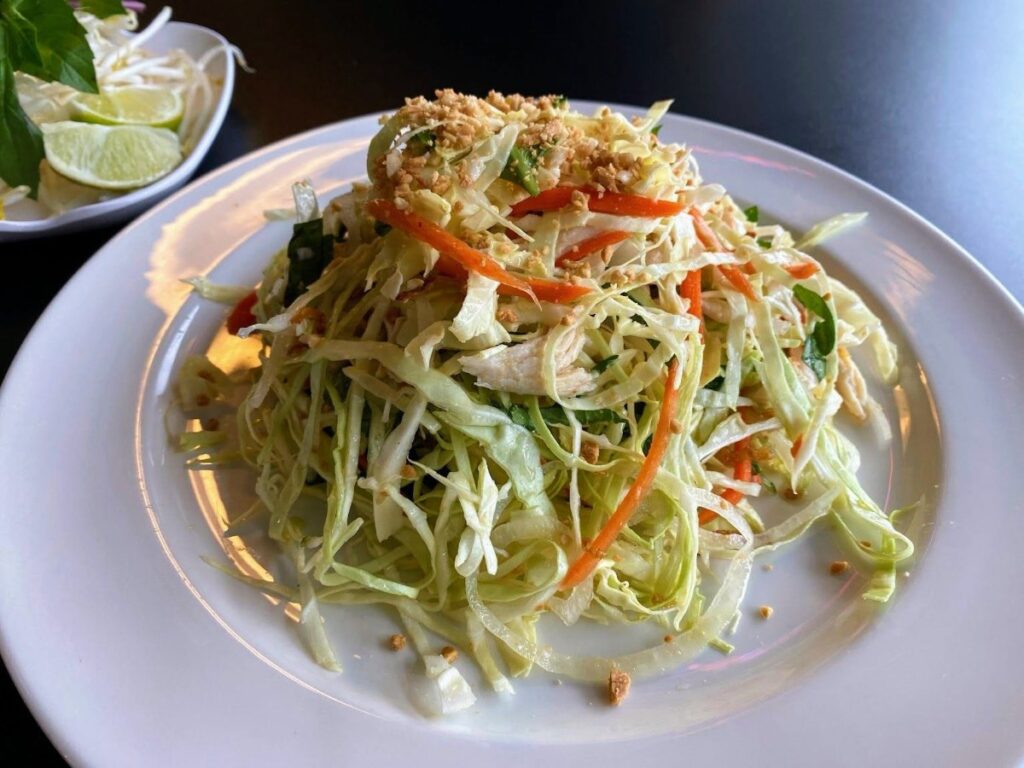 Shredded cabbage salad with carrot ribbons and crushed peanuts on a white plate, lime wedges visible in the background plate garnish nearby.