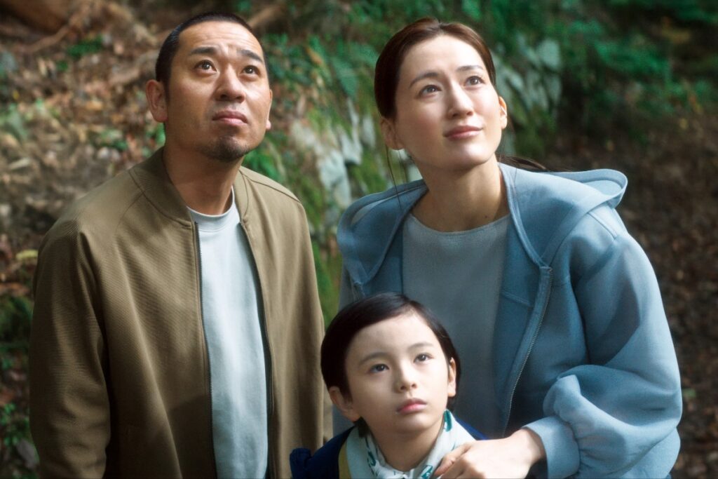 A family of three — man, woman, and young girl — gaze upward in a forested outdoor setting with greenery behind them.