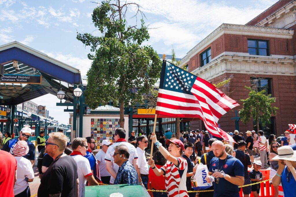 sound-transit-world-cup-3 - junglecity.com Crowd of people at an outdoor street event, a large American flag waved in the foreground with a brick building and train station signage in the background.